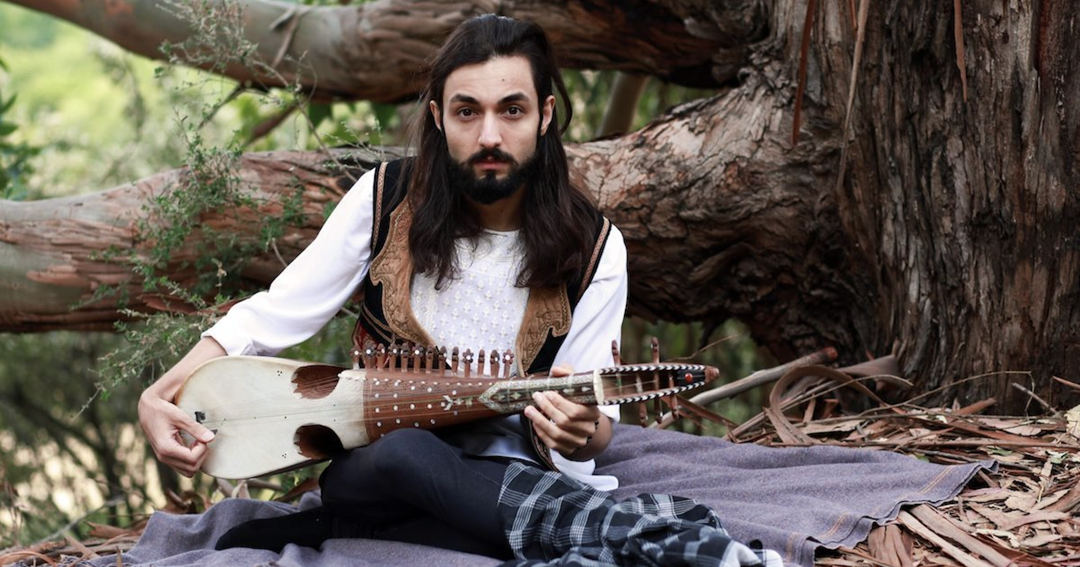 Qais Essar poses with his rabab, seated on a blanked by a tree in the forest